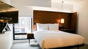 Una habitación de hotel moderna con una cama grande, un cabecero de madera, una mesita de noche, una lámpara y vistas a un edificio a través de una ventana.