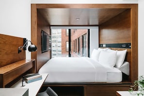 A modern hotel room with a wooden headboard, a large bed with white bedding, a desk with a lamp, and a view of a brick building through the window.