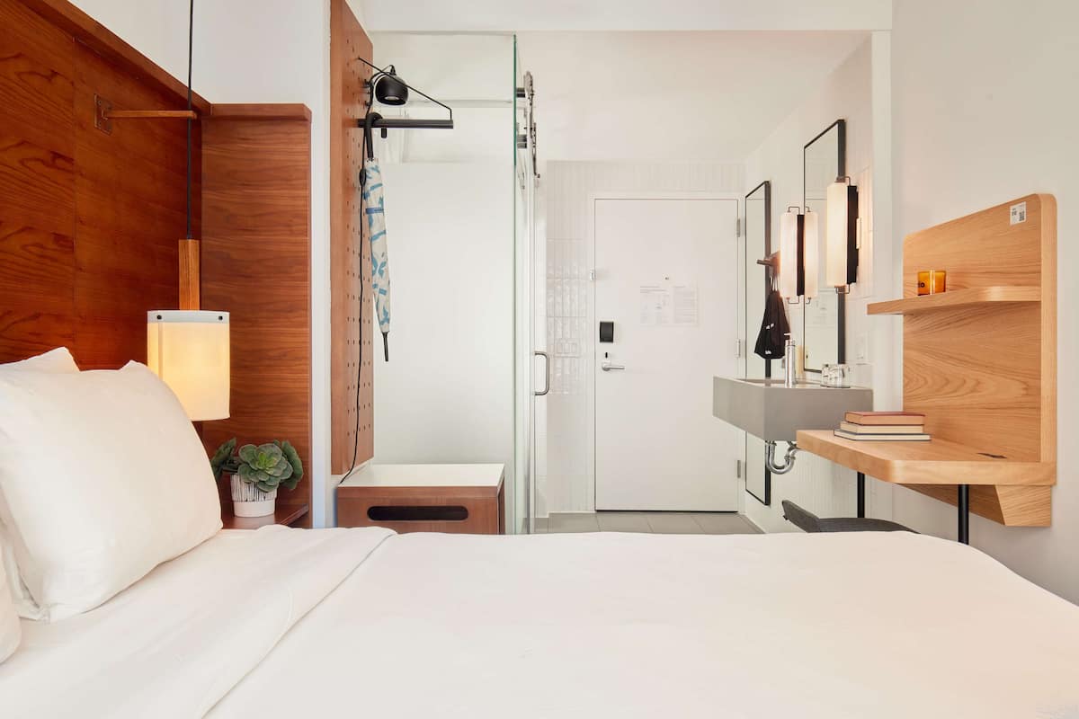A modern hotel room with a wooden headboard, a bedside lamp, a small plant, a white door, a mirror, and a wooden shelf with books.