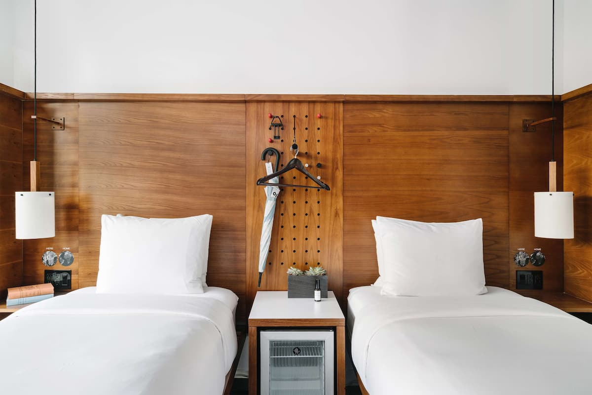 A hotel room with two beds, wooden headboard, bedside tables with lamps, a small heater, and a wall-mounted coat rack.
