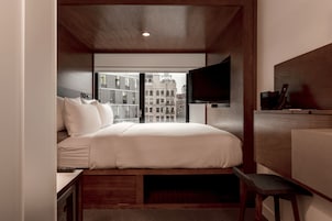 A compact hotel room with a bed, a TV mounted on the wall, a small desk, and a view of the cityscape through the window.