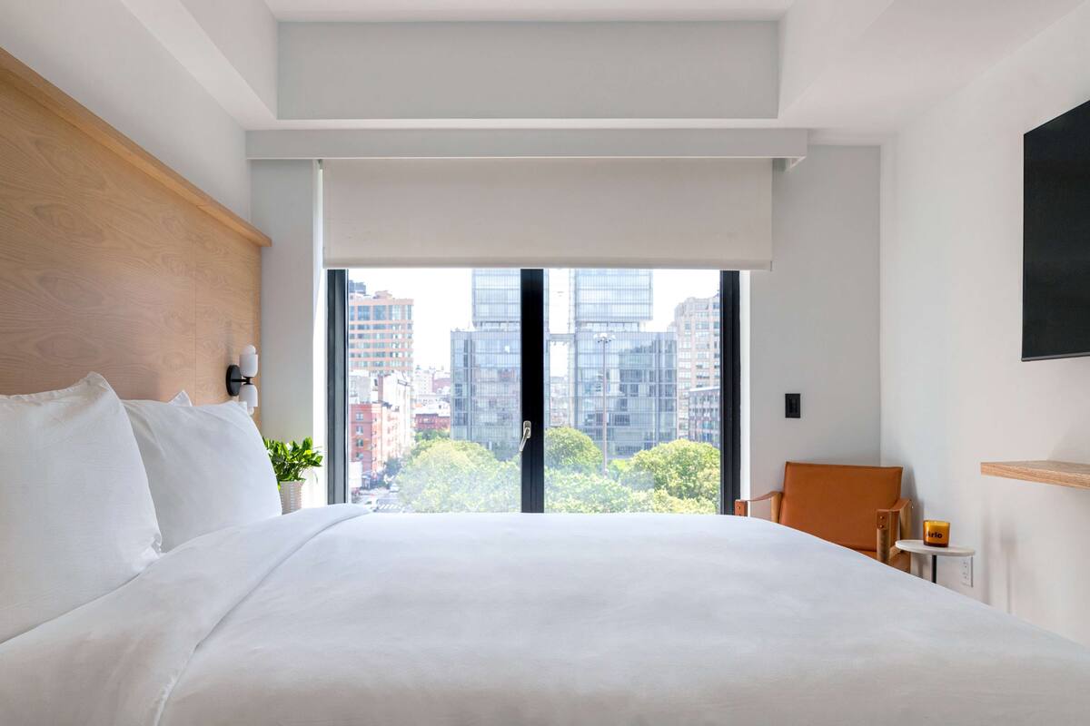 A modern hotel room with a large bed, a view of city buildings, a wooden headboard, and a small table with a plant.