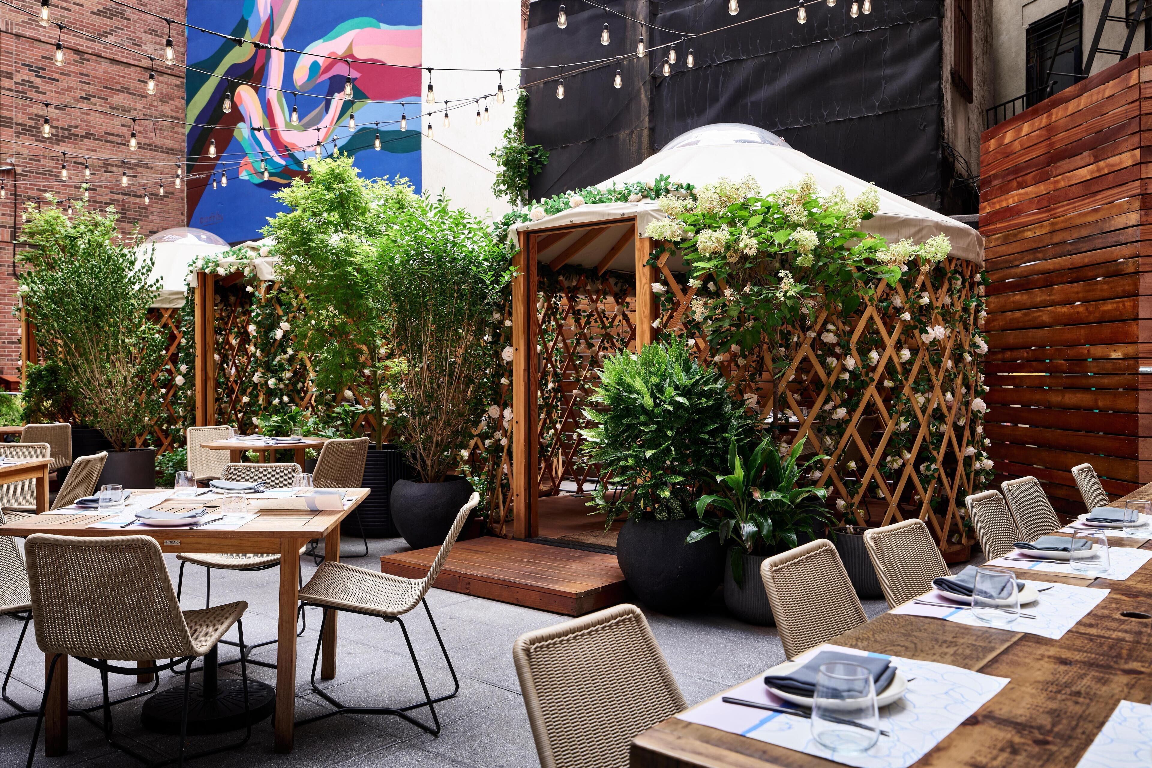An outdoor dining area with wooden tables and wicker chairs, surrounded by potted plants and a mural on the wall.