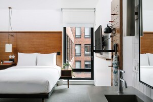 Quarto de hotel moderno com uma cama grande, uma escrivaninha, uma luminária de mesa e vista para um edifício de tijolos pela janela.