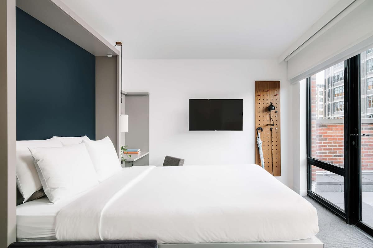 A modern hotel room with a large bed, a desk with a chair, a flat-screen TV mounted on the wall, and a large window offering a city view.
