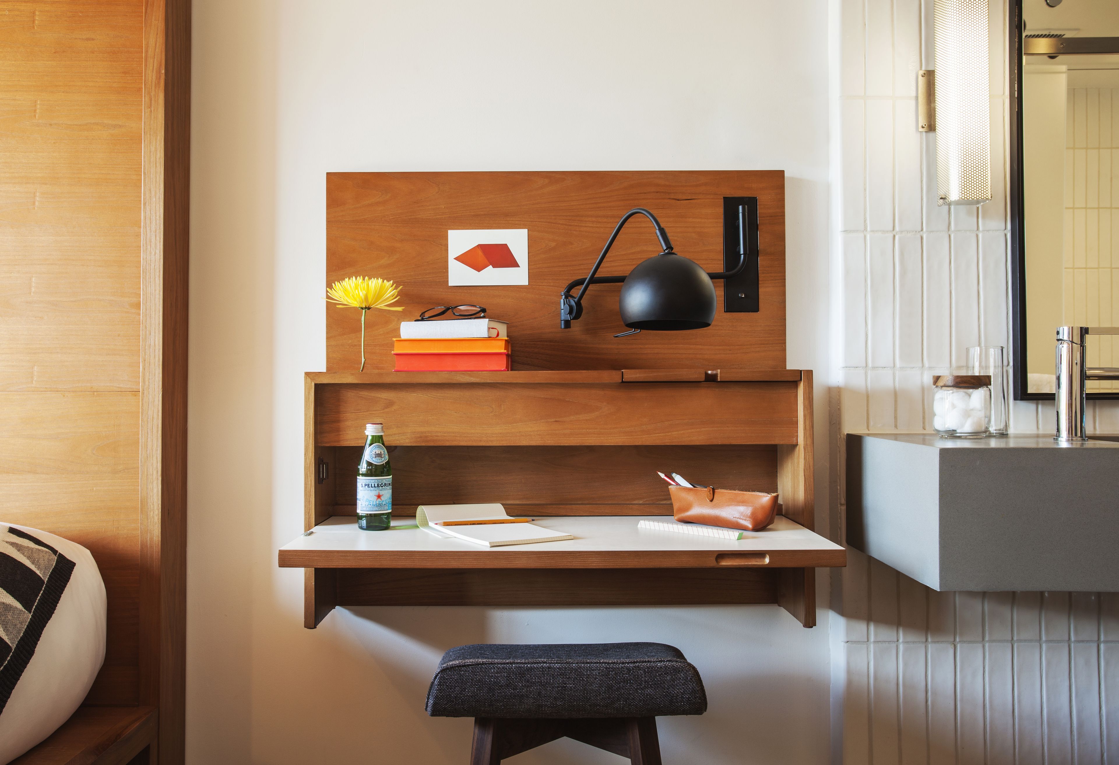 A modern workspace with a wooden desk, a black desk lamp, a notebook, a pen, a bottle of water, and a small stool.