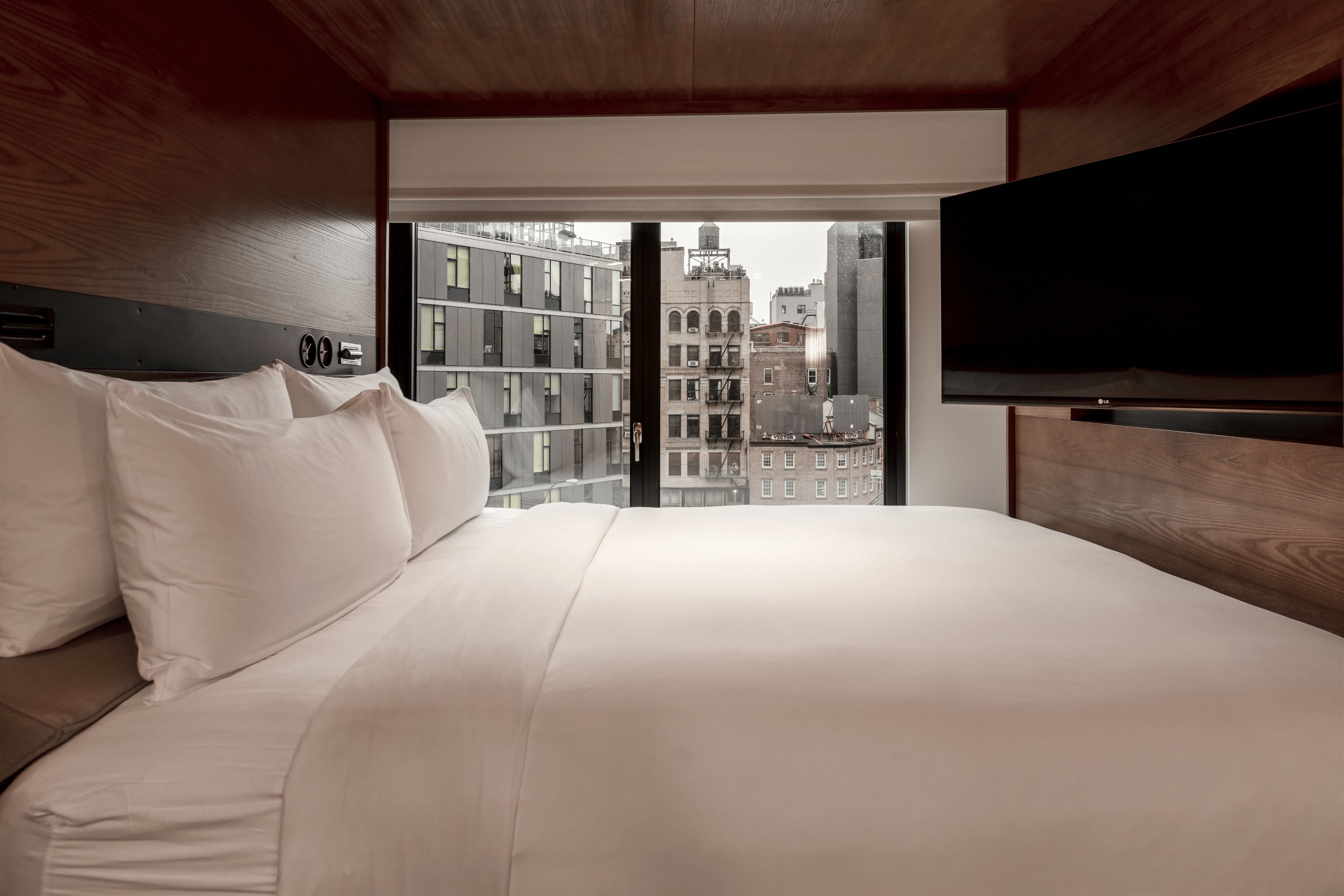 A hotel room with a large bed, a flat-screen TV, and a view of city buildings through a large window.