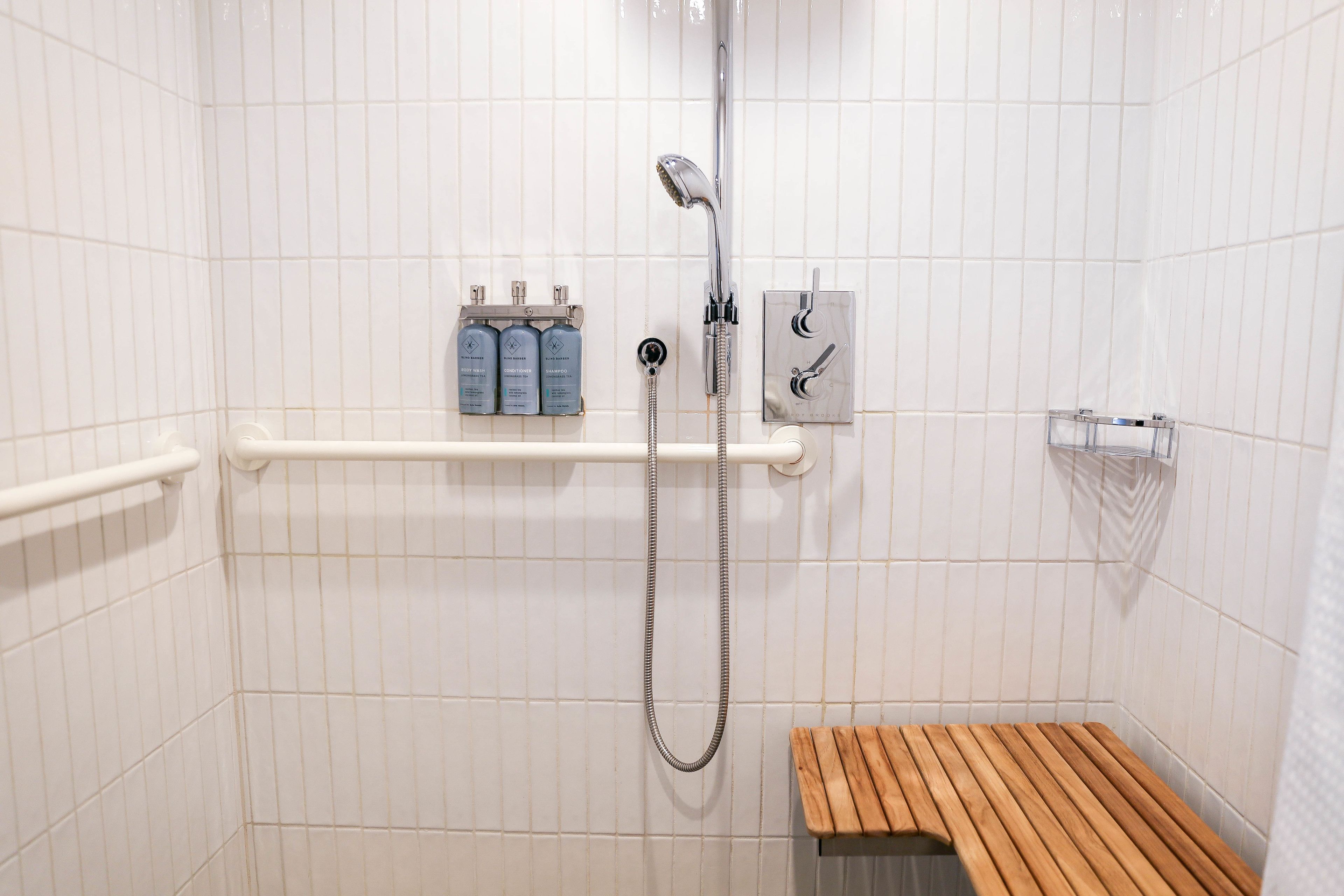 A shower area with white tiled walls, a wooden bench, a handheld showerhead, and a soap dispenser on a shelf.
