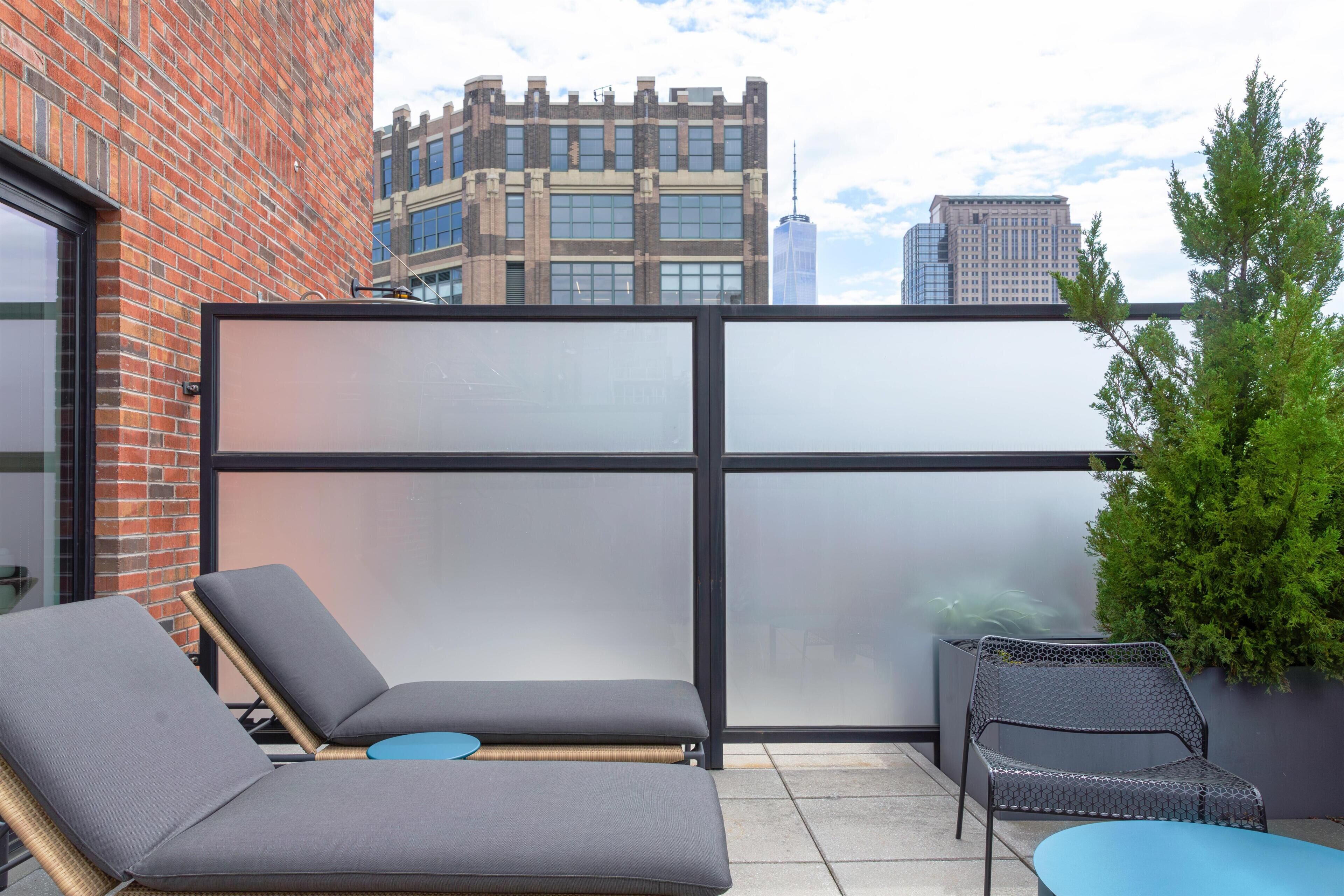 A rooftop terrace with two lounge chairs and a small table, a glass partition, and a view of city buildings.