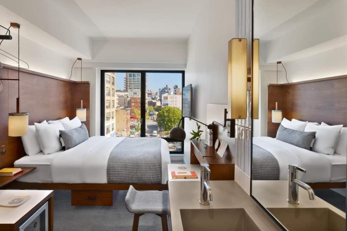 A hotel room with a large bed, a desk, a chair, a mirror, a sink, and a view of the city.