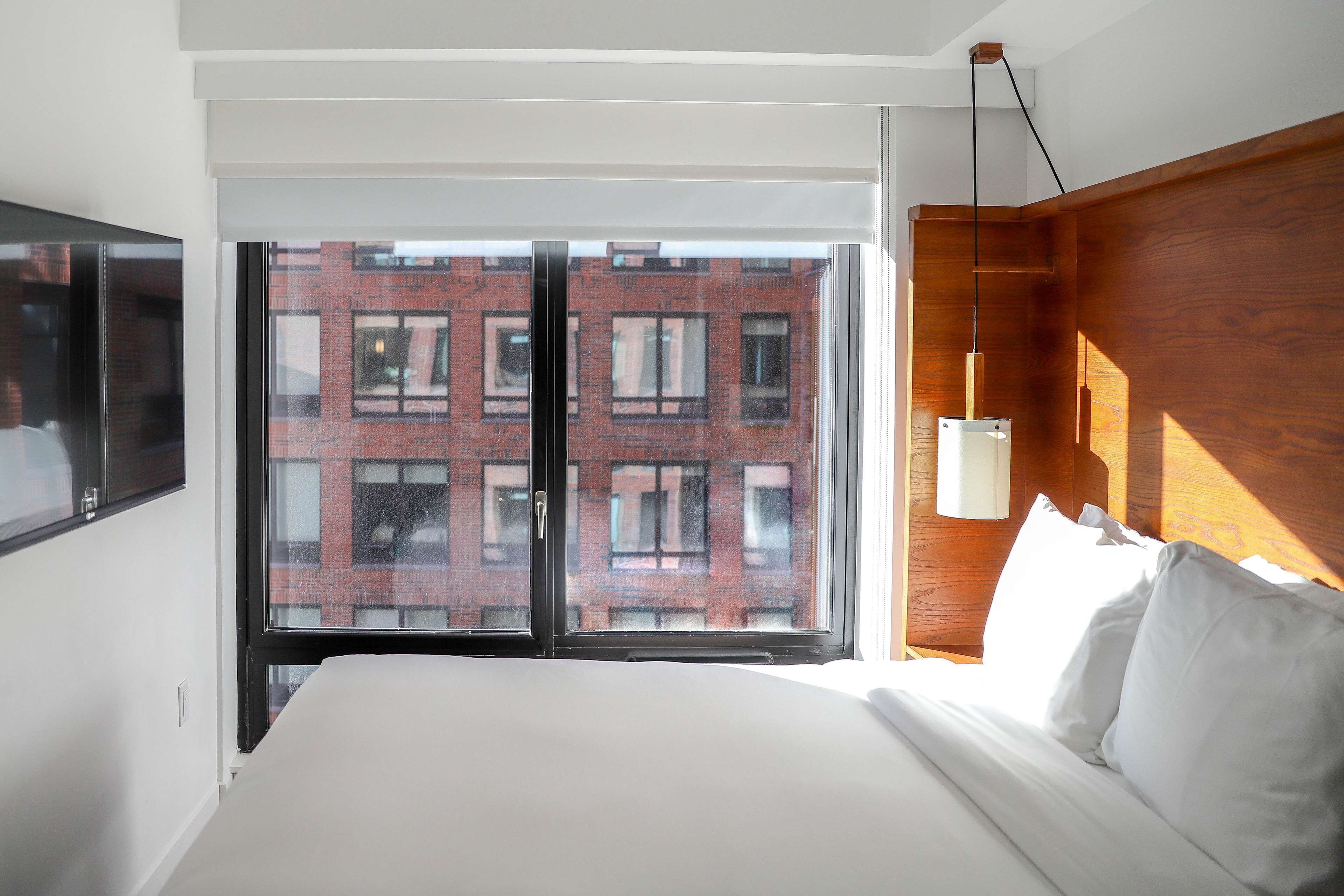 A hotel room with a large bed, a wooden headboard, a window with a view of a brick building, and a wall-mounted TV.