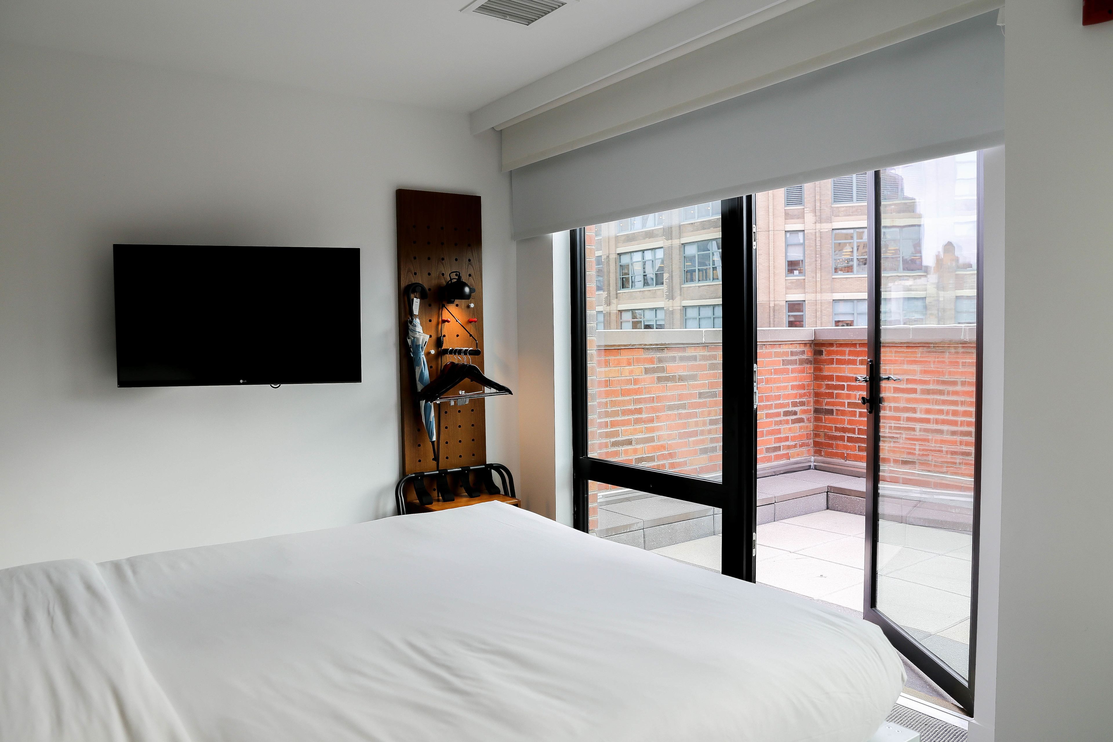 Una habitación de hotel moderna con una cama grande, un televisor montado, un panel de pared de madera y vistas a un edificio de ladrillo a través de una puerta corrediza de vidrio.
