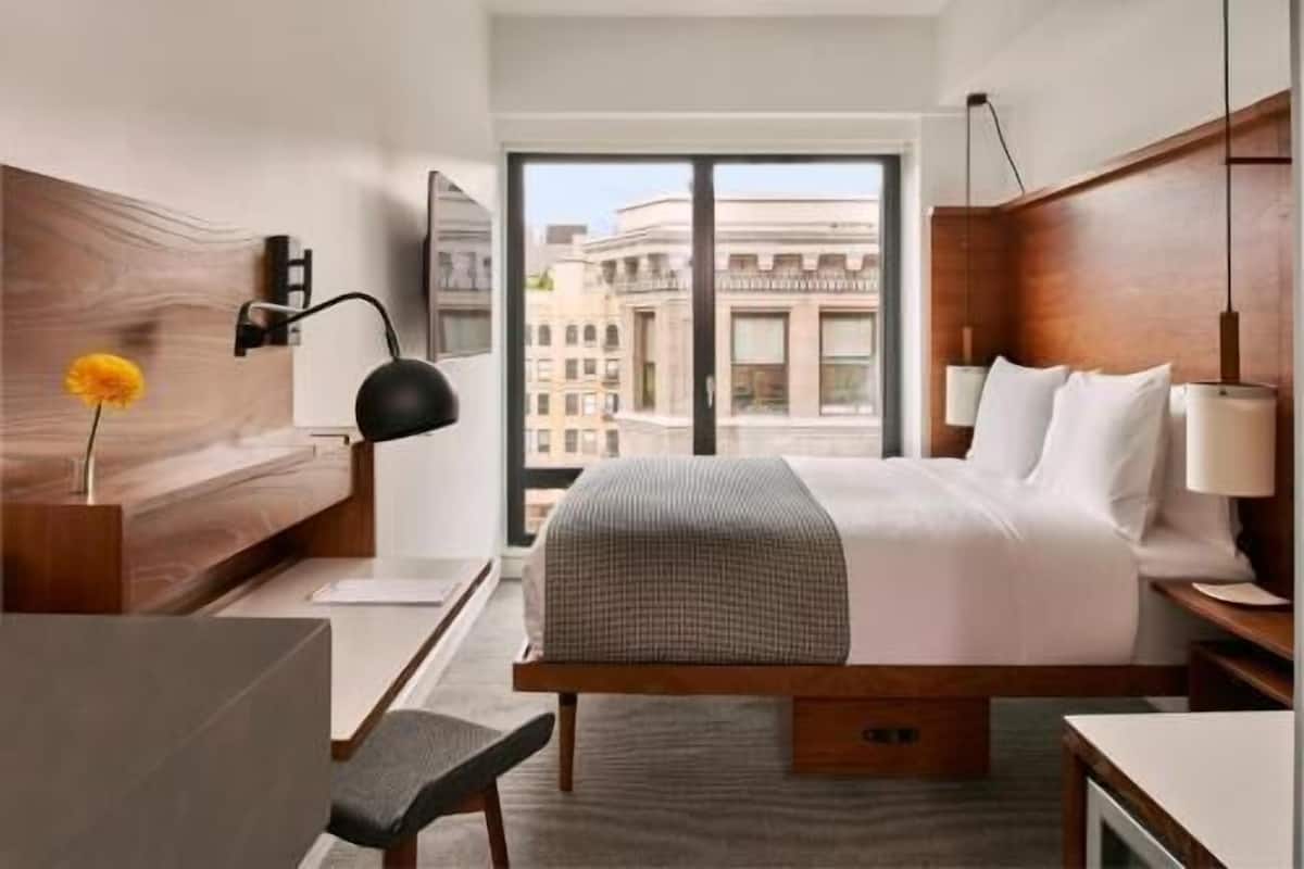 A hotel room with a large bed, a desk, a chair, a lamp, and a view of buildings through the window.