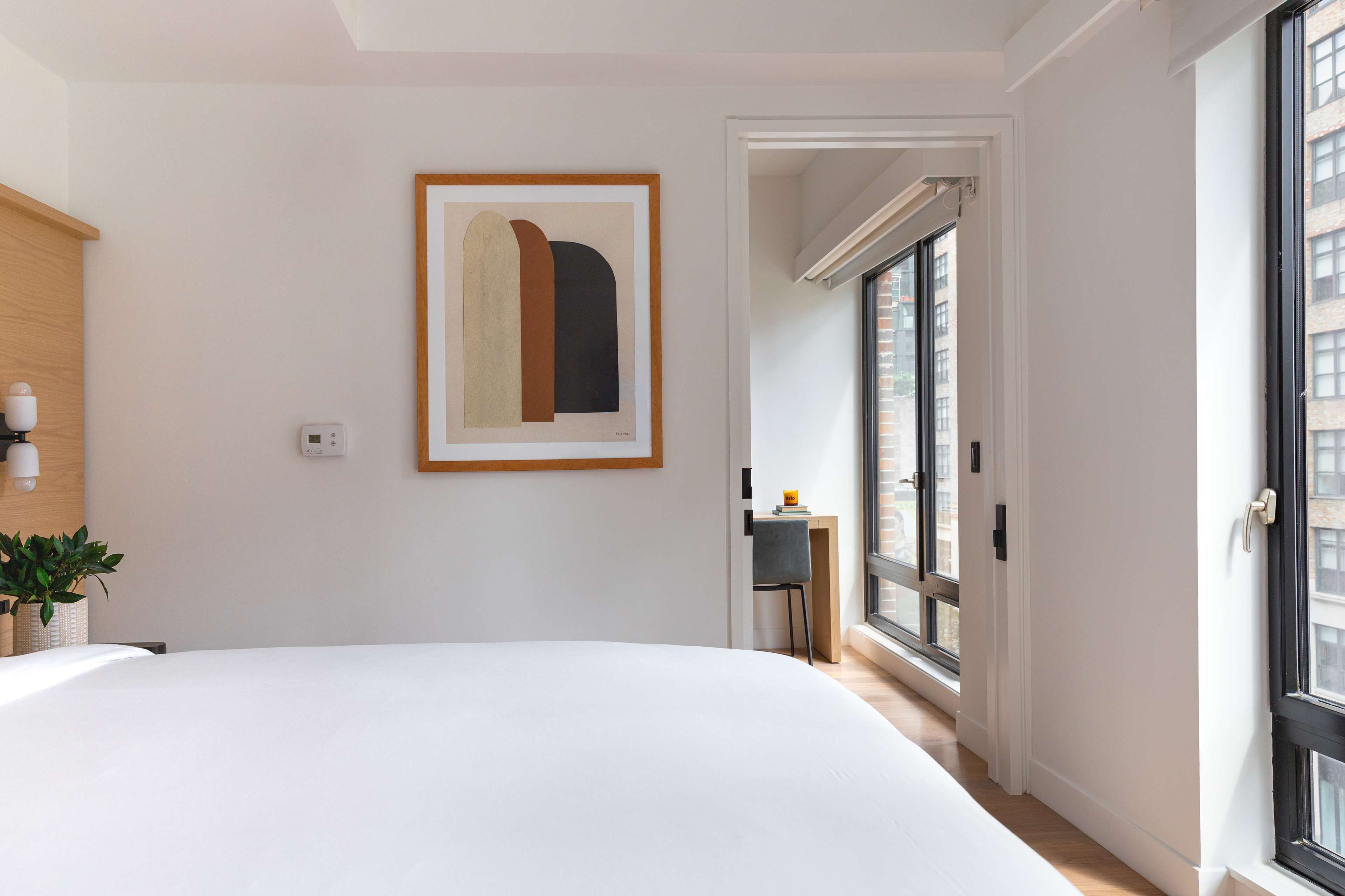 Une chambre d’hôtel moderne dotée d’un grand lit, d’une œuvre d’art abstraite encadrée, d’un petit bureau avec une chaise et offrant une vue sur la ville grâce à de grandes fenêtres.