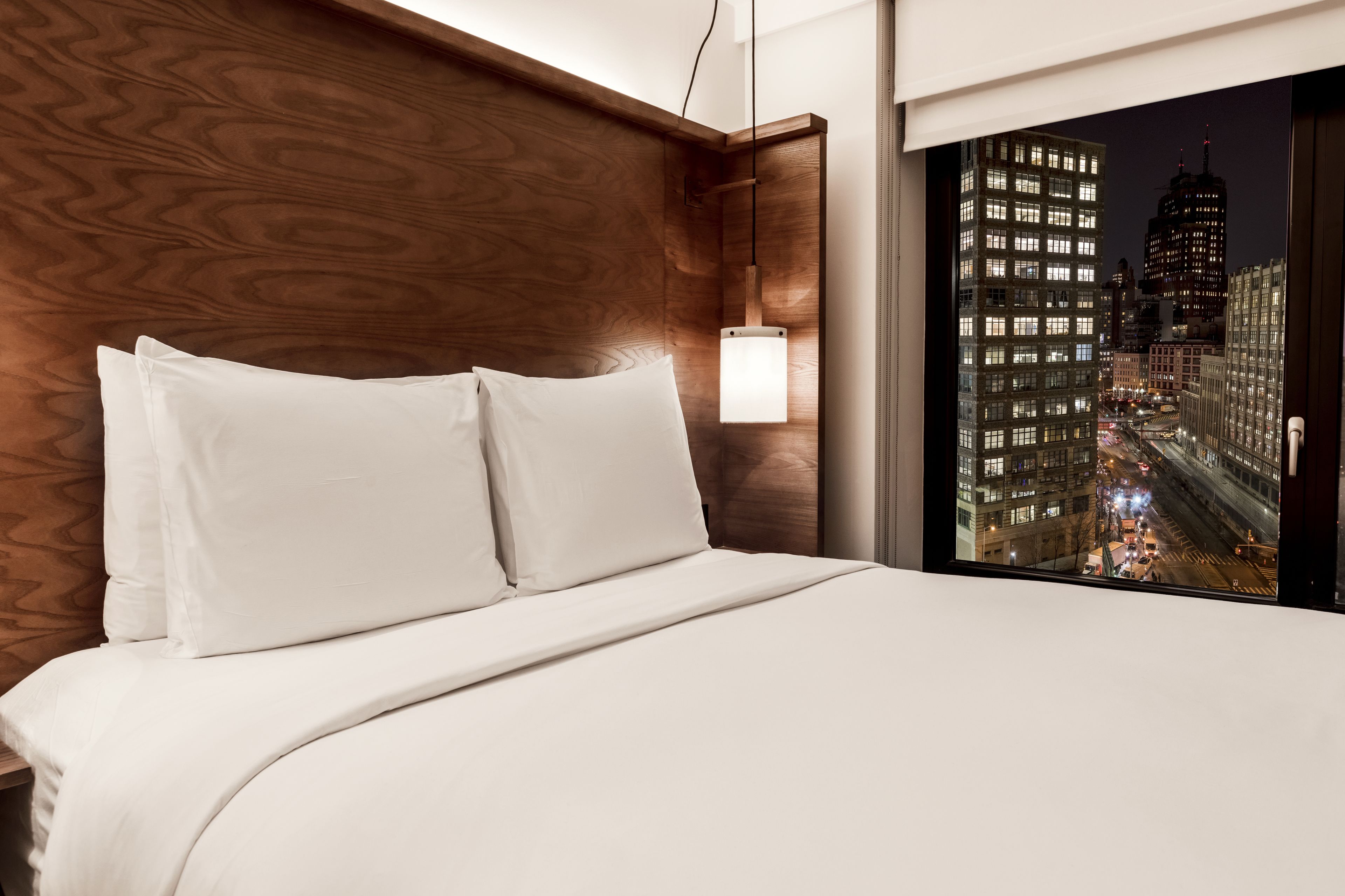 A hotel room with a large bed, wooden headboard, and a view of a cityscape at night.