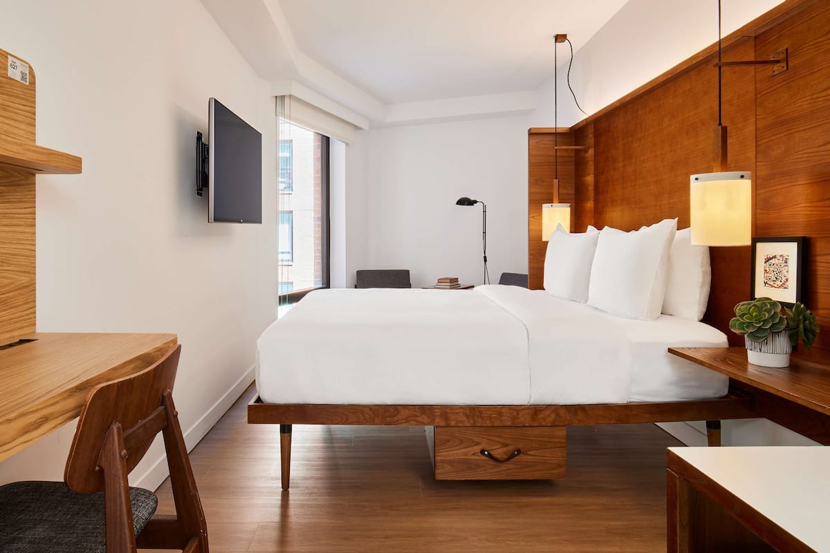 A modern hotel room with a wooden bed frame, a flat-screen TV mounted on the wall, a desk with a chair, and a bedside table with a lamp and a plant.