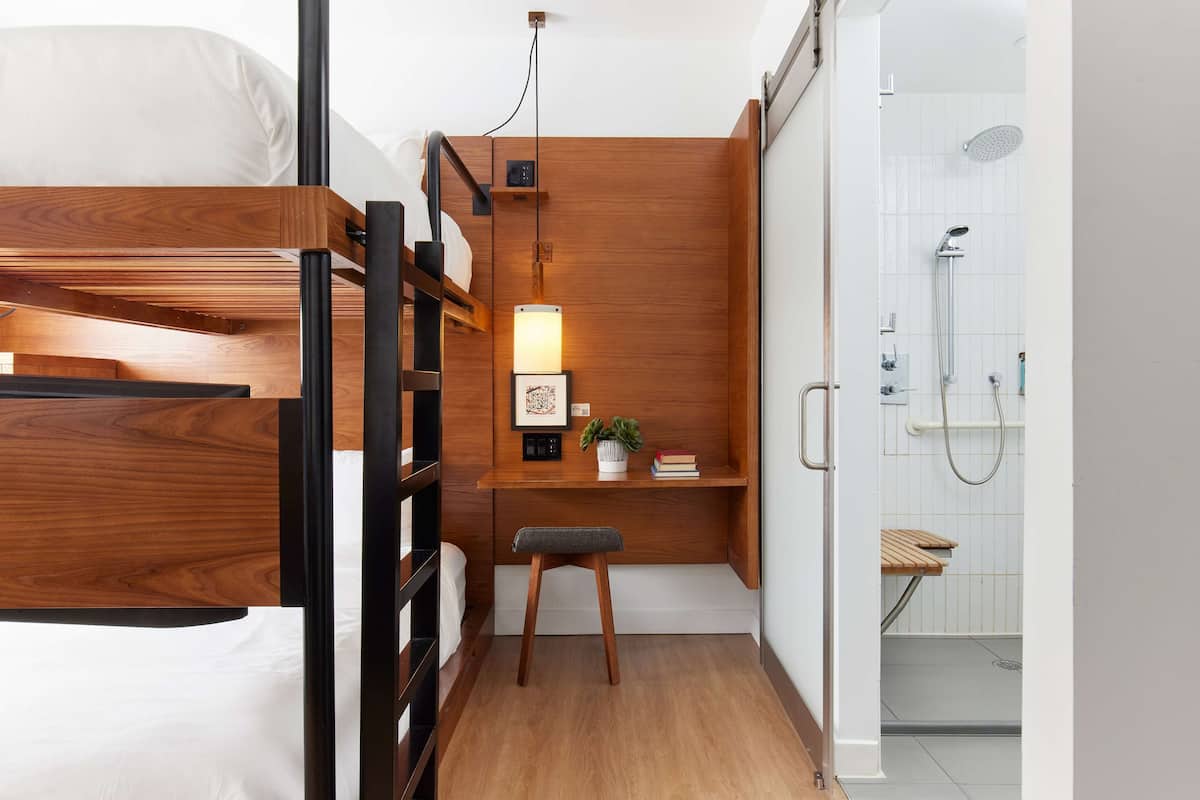 A compact room with a bunk bed, a desk with a chair, a small plant, and a shower area.