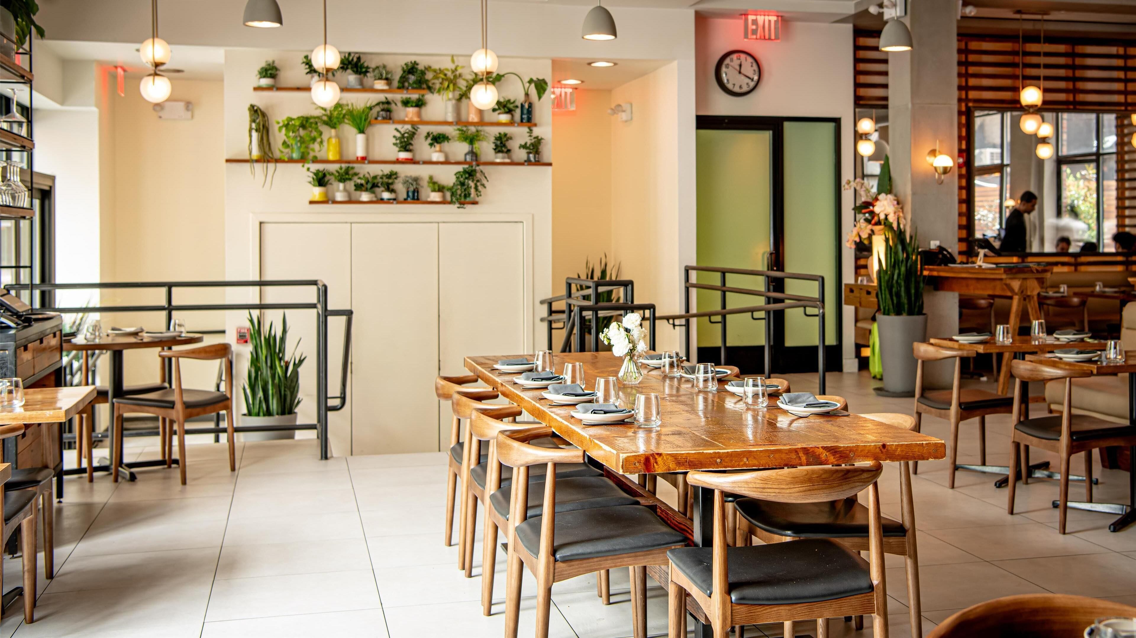 Een restaurant met houten tafels en stoelen, plantenpotten op planken en een vluchtroutebord.