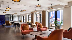A spacious indoor area with multiple seating arrangements, including round tables and cushioned chairs, and large windows providing views of the outside. - Arlo SoHo (New York)