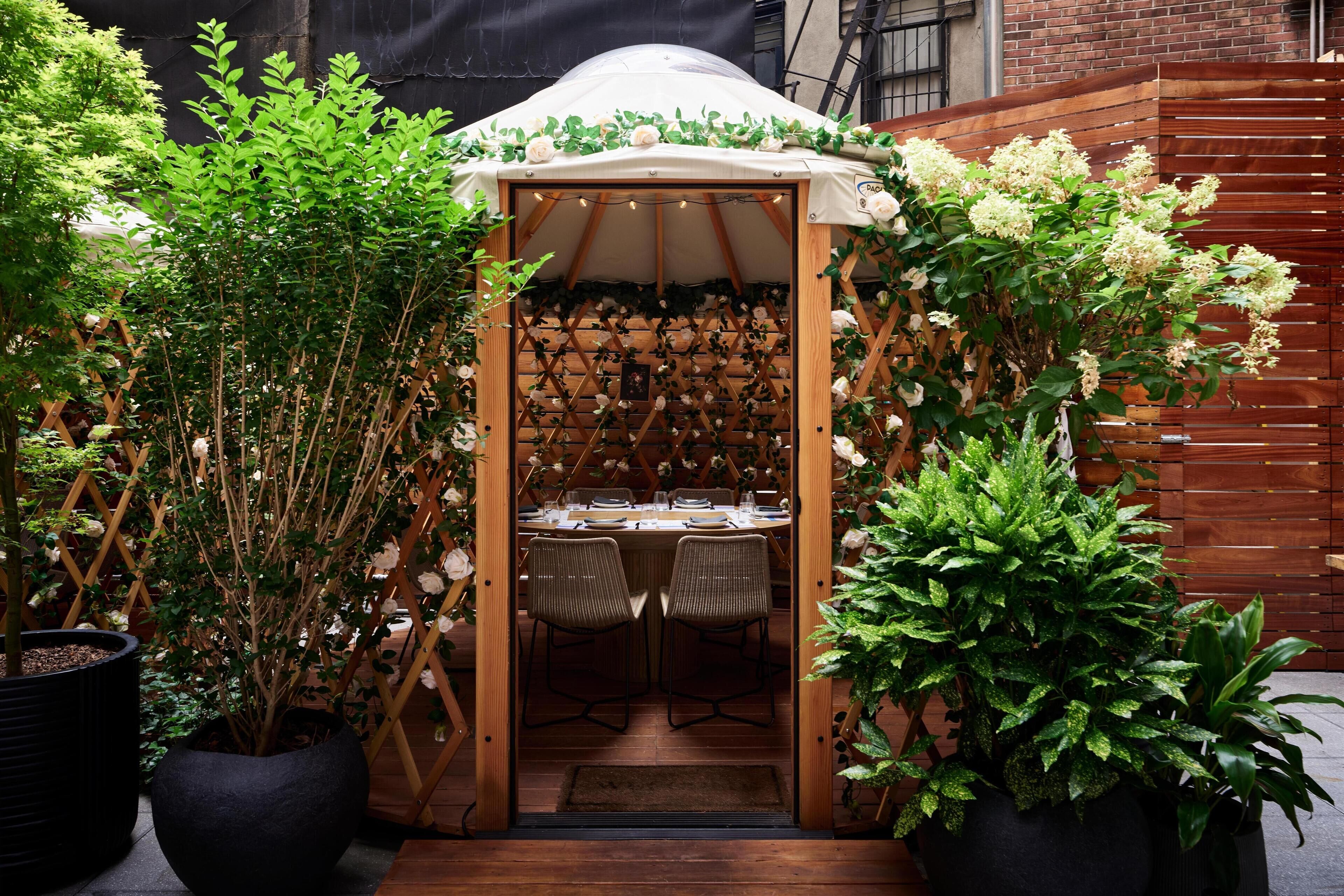 Un petit espace extérieur aménagé pour les repas, à l’abri, avec une table et des chaises en bois, entouré de plantes en pot et d’une clôture en bois.