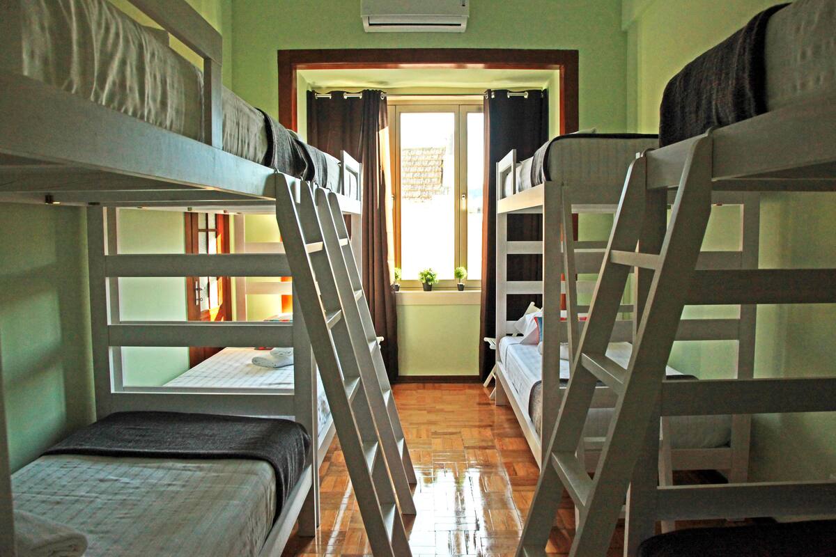 A dormitory room with bunk beds, a window with curtains, and a small table with a lamp.