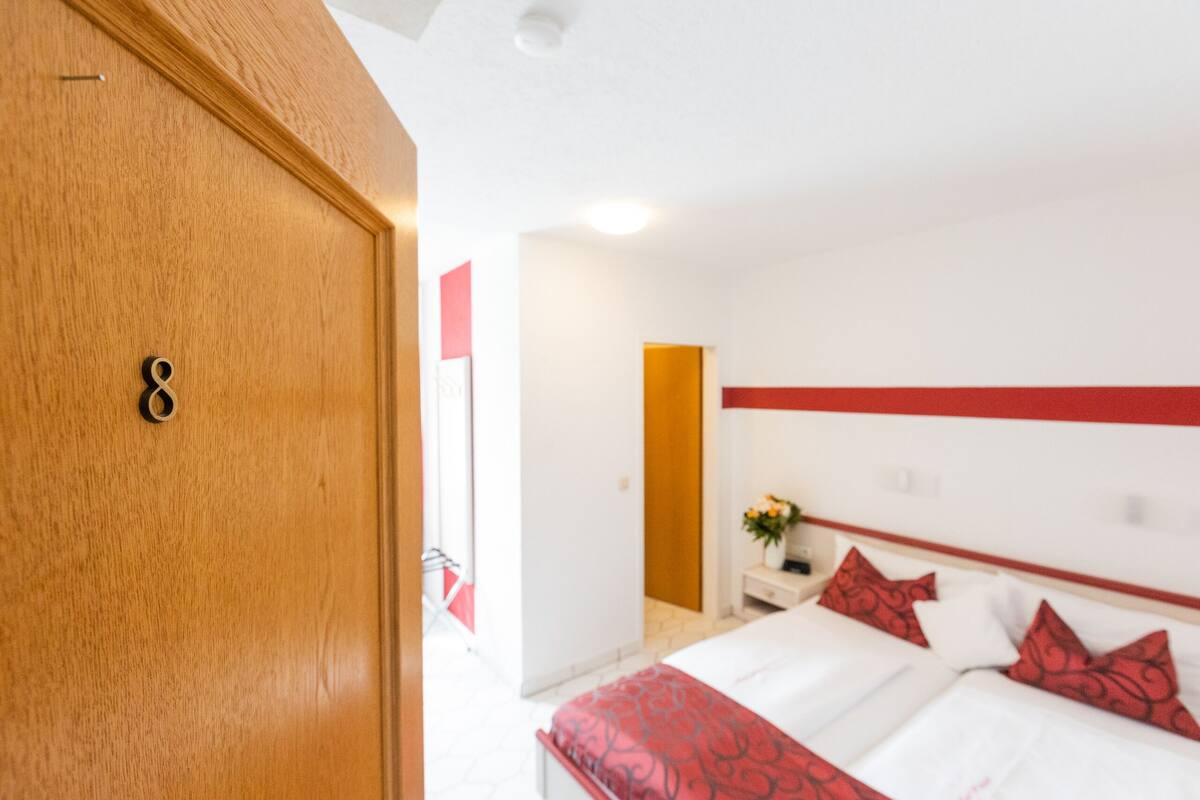 Ein Hotelzimmer mit Holztür, einem Bett mit rotem und weißem Bettzeug, einem Nachttisch mit Blumenvase und einer Wandgestaltung in Rot und Weiß.