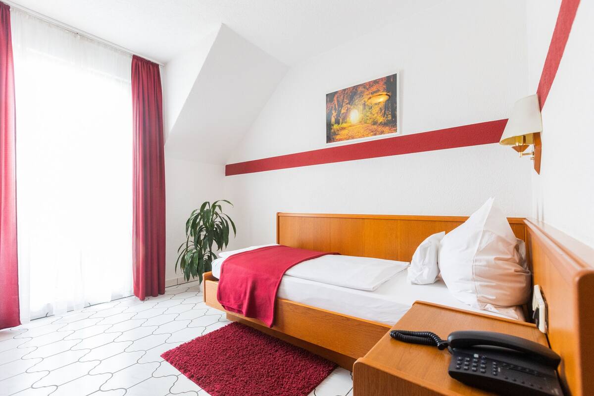 Ein Hotelzimmer mit einem hölzernen Bett, einer roten Tagesdecke, einem weißen Kopfkissen, einer roten Akzentwand, einem Gemälde, einer Pflanze und einem Telefon auf einem Nachttisch aus Holz.