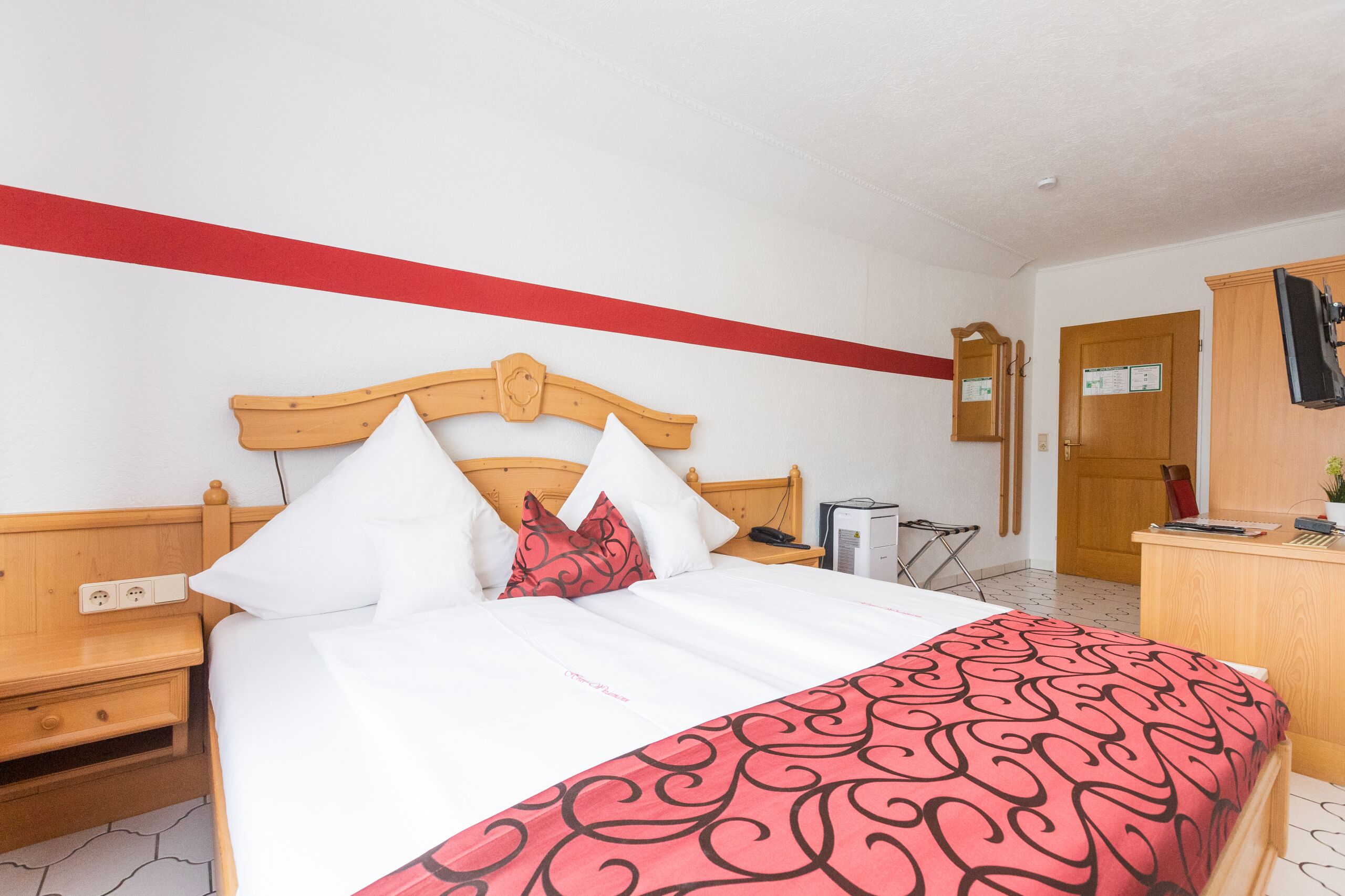 A hotel room with a wooden bed, white bedding, a red and black patterned bedspread, a wooden nightstand, a desk with a computer, and a mirror on the wall.