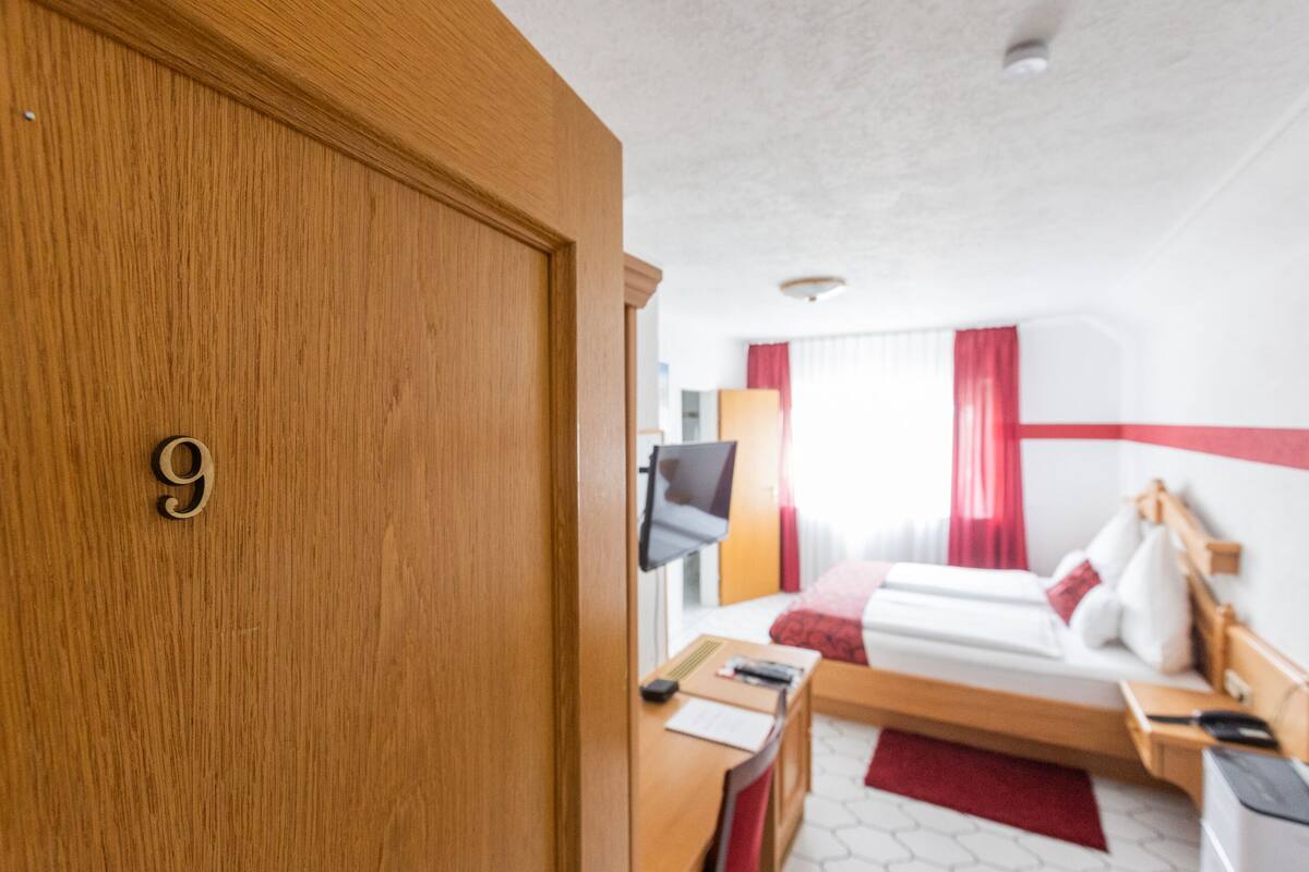 Ein Hotelzimmer mit einer Holztür, die mit der Nummer 9 gekennzeichnet ist, einem Bett mit weißer und roter Bettwäsche, einem an der Wand befestigten Fernseher und einem Schreibtisch mit rotem Teppich.
