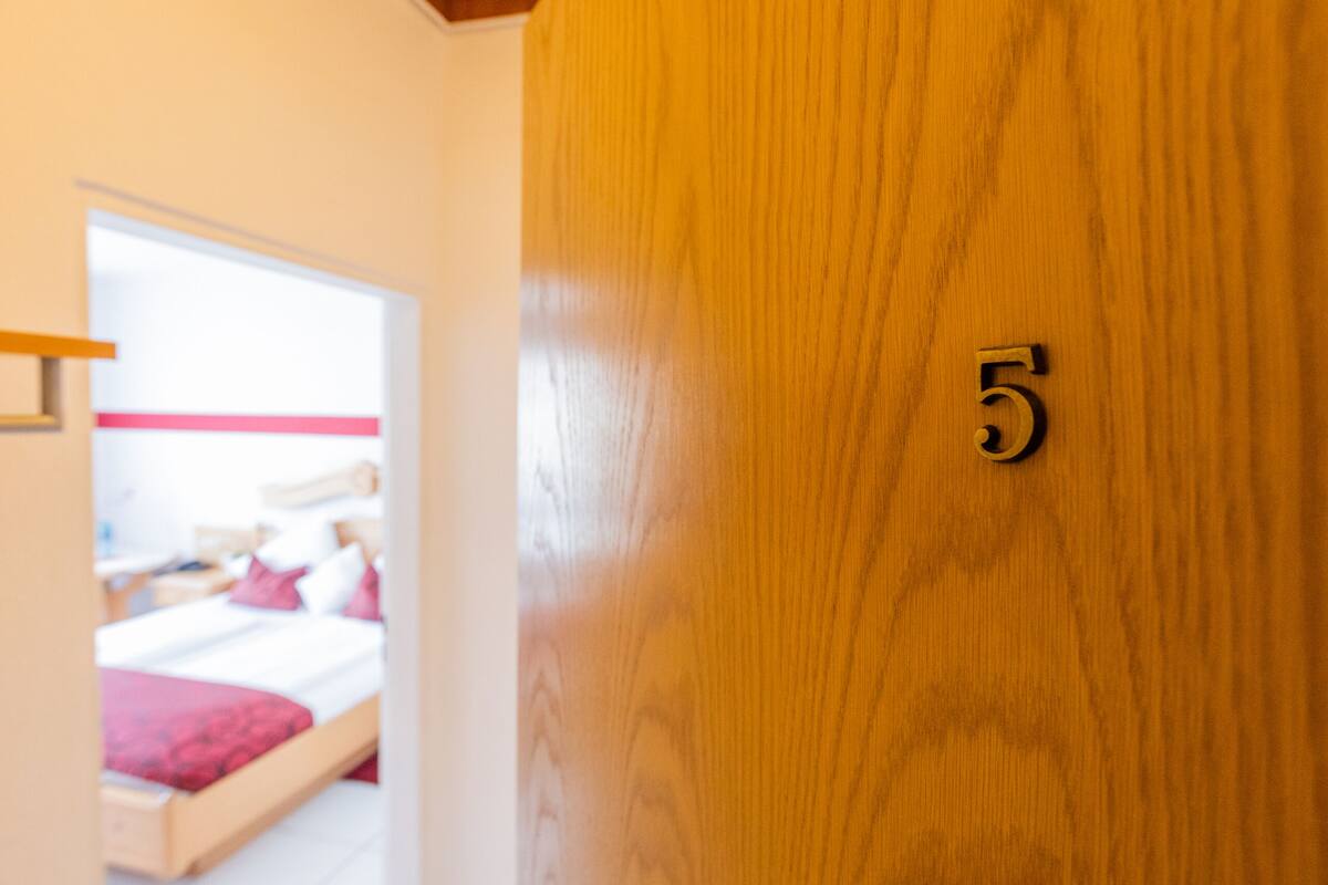 Eine Holztür mit der Nummer 5, die zu einem Raum mit einem Bett und einer roten Decke führt.