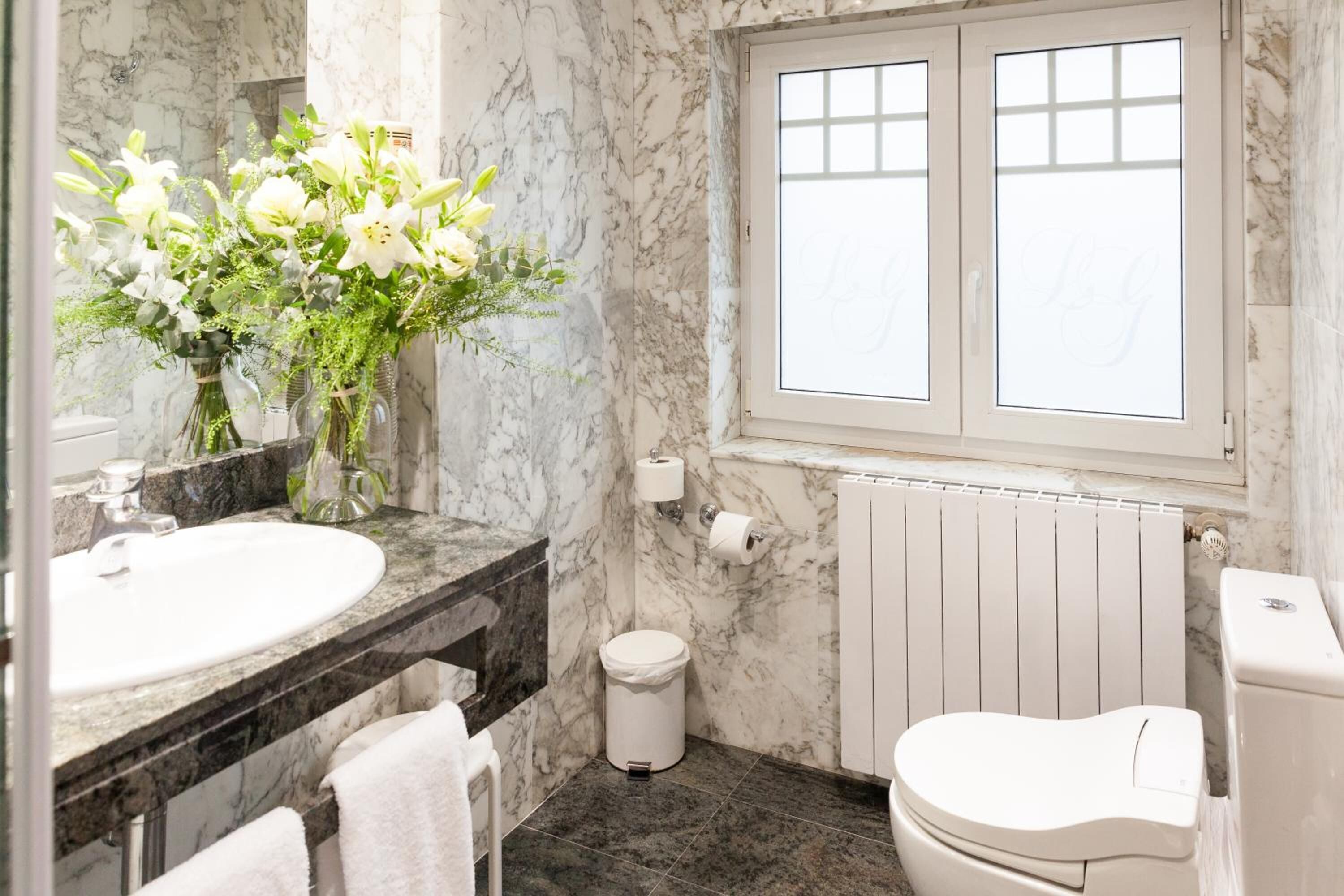 Une salle de bain avec un mur en marbre, des toilettes blanches, un lavabo blanc, un miroir et une fenêtre en verre dépoli.