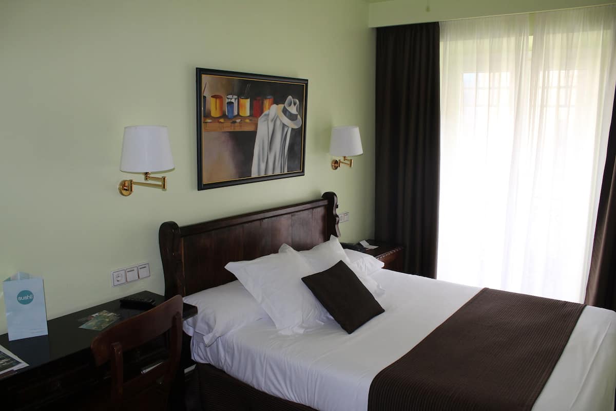 Ein Hotelzimmer mit einem großen Bett, zwei Nachttischen, einem Schreibtisch, einem Stuhl, einem Bild an der Wand und einem Fenster mit Vorhängen.