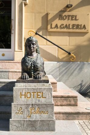A statue of a seated figure with a plaque reading "HOTEL LA GALERIA" in front of a building with a similar sign.