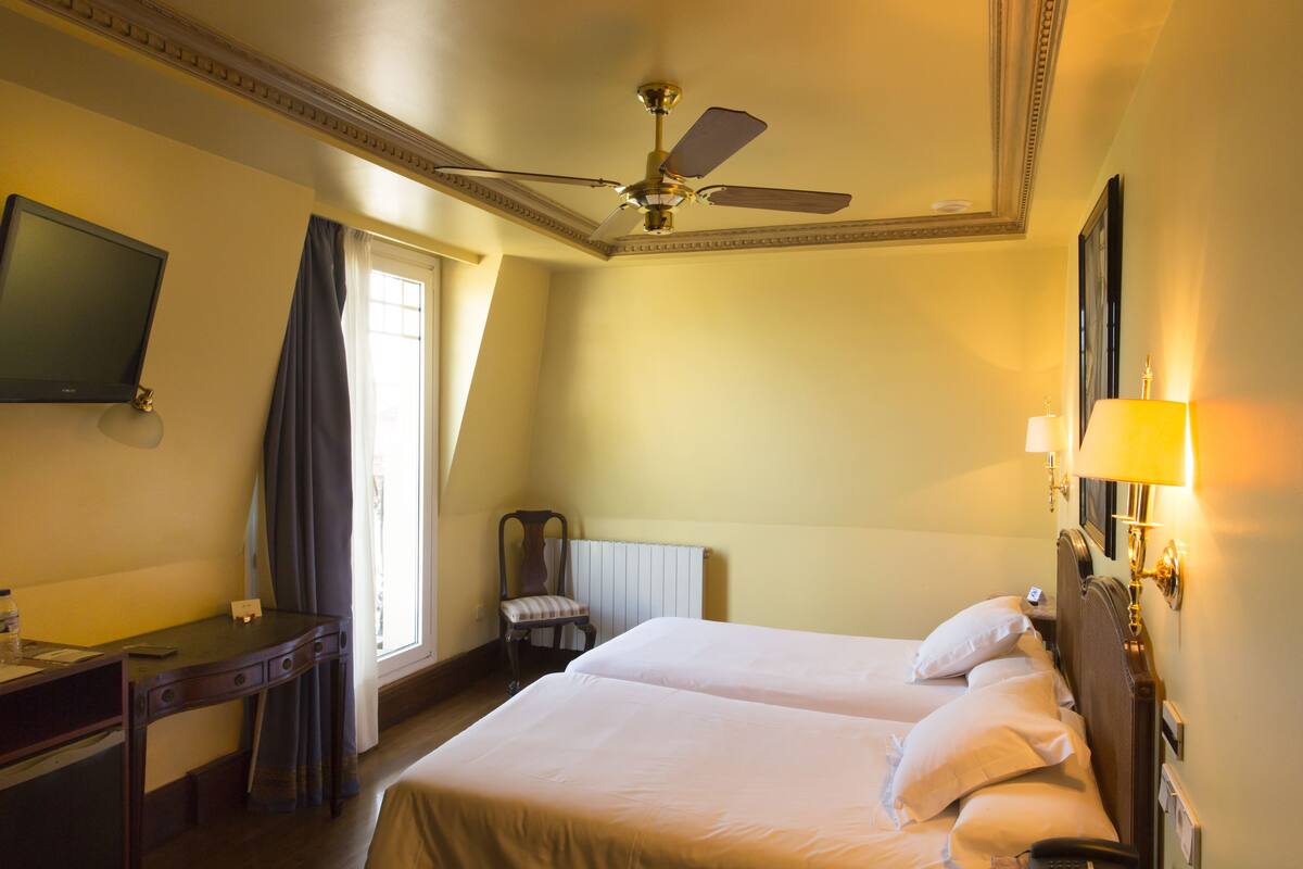 Ein Hotelzimmer mit zwei Betten, einem Schreibtisch, einem Stuhl, einem Deckenventilator, einem Fernseher und einem Fenster mit Vorhängen.