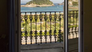 A room with a large sliding glass door leading to a balcony with a view of the sea and a hill. - Hotel La Galería (San Sebastián)