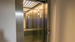 Interior de un ascensor con paredes metálicas, un panel de control y un techo con una rejilla de luces.