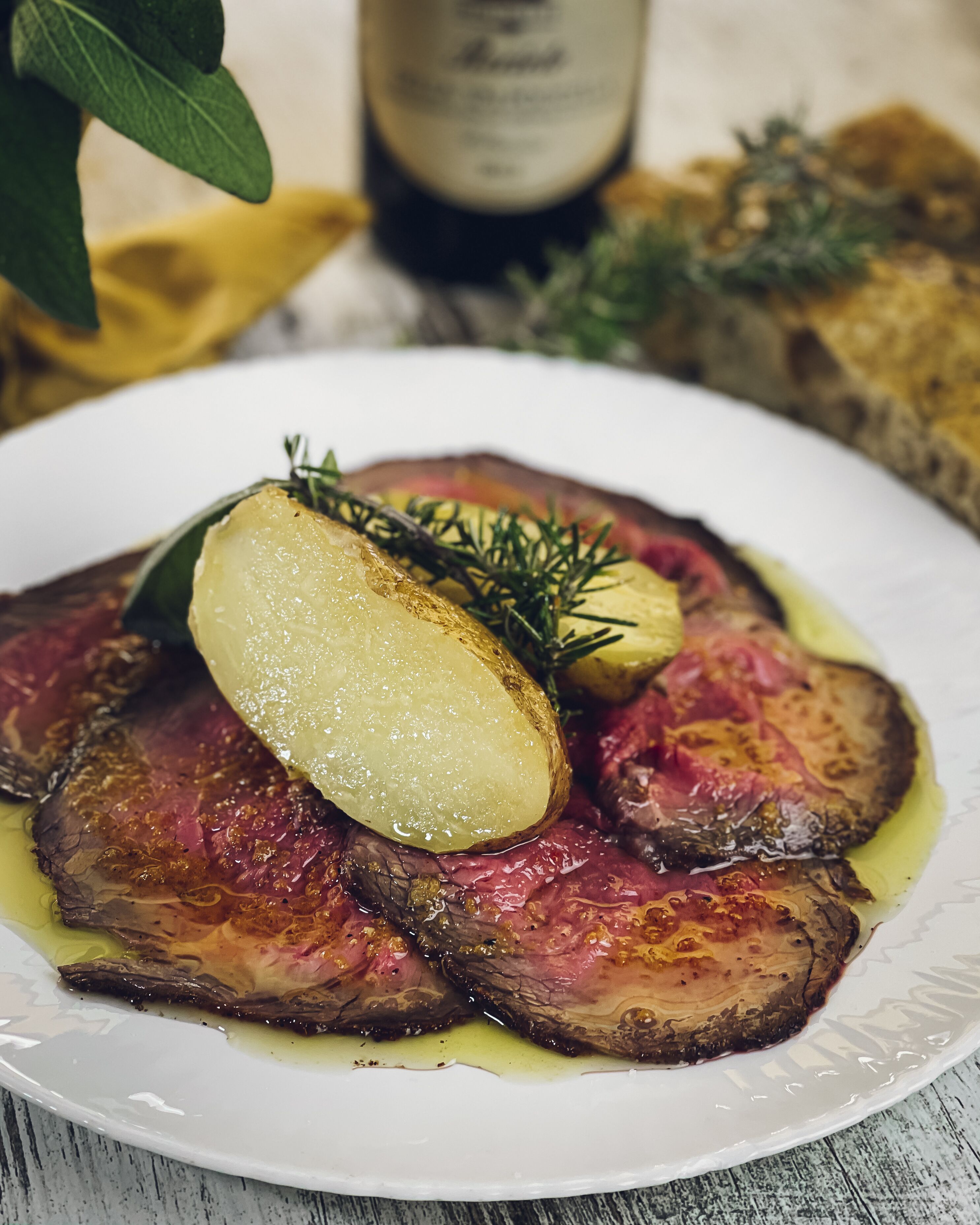 Un plato de carne con una rodaja de patata y una ramita de romero.