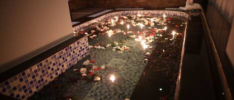 Una bañera llena de pétalos de rosa y velas, con un muro de azulejos y una pared de ladrillo al fondo.