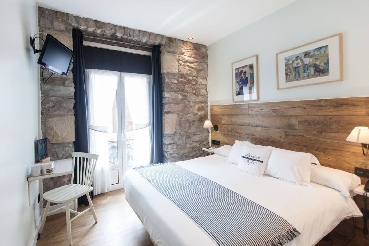 A hotel room with a stone wall, a bed with white linens, a wooden headboard, a desk with a chair, and a TV mounted on the wall.