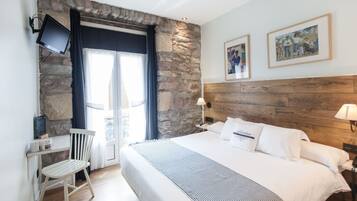 Habitación de hotel con pared de piedra, cama con sábanas blancas, cabecero de madera, escritorio con silla y televisor montado en la pared.