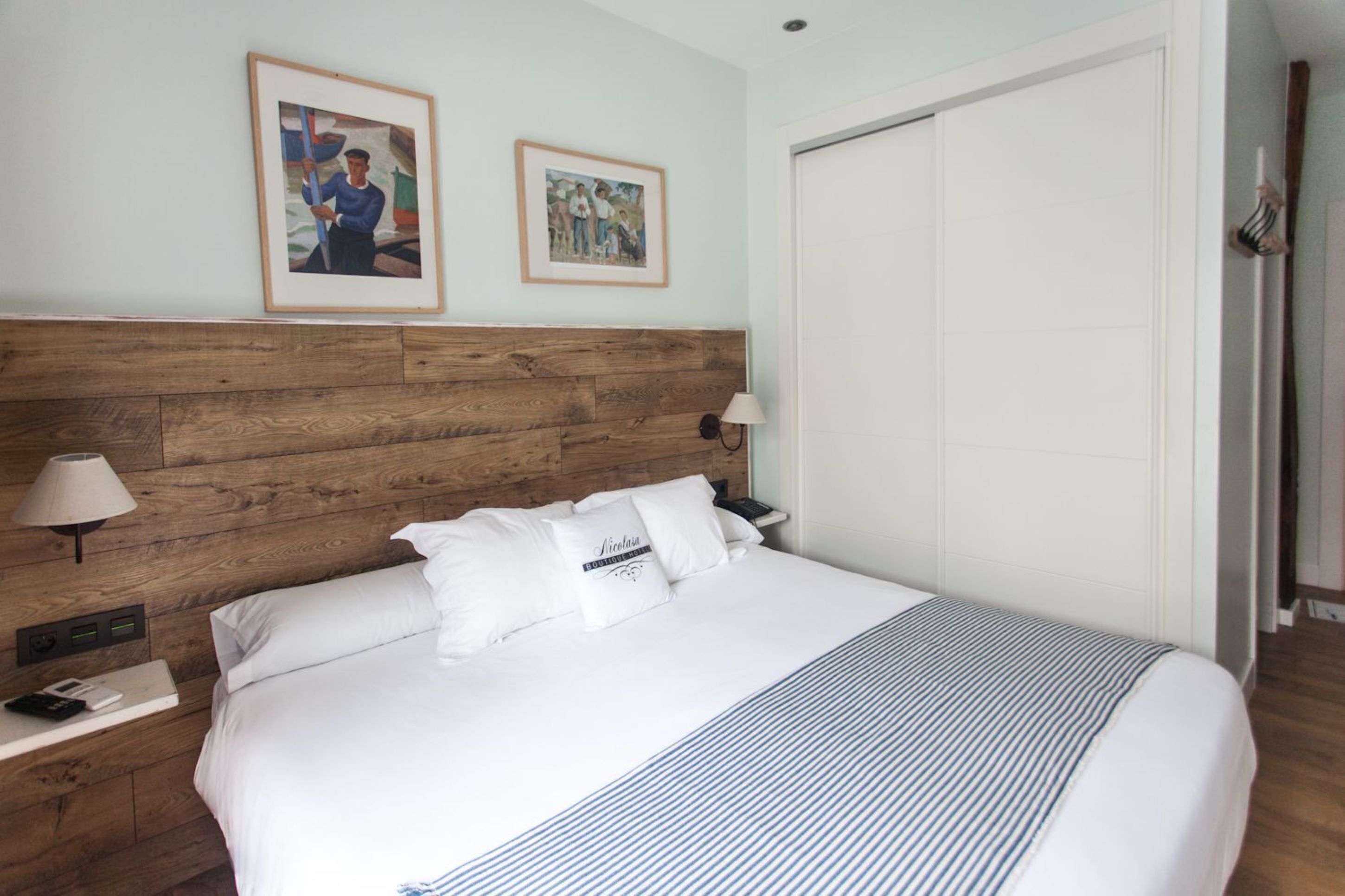 A hotel room with a wooden headboard, white bedding, two framed pictures on the wall, a bedside lamp, and a closed door.
