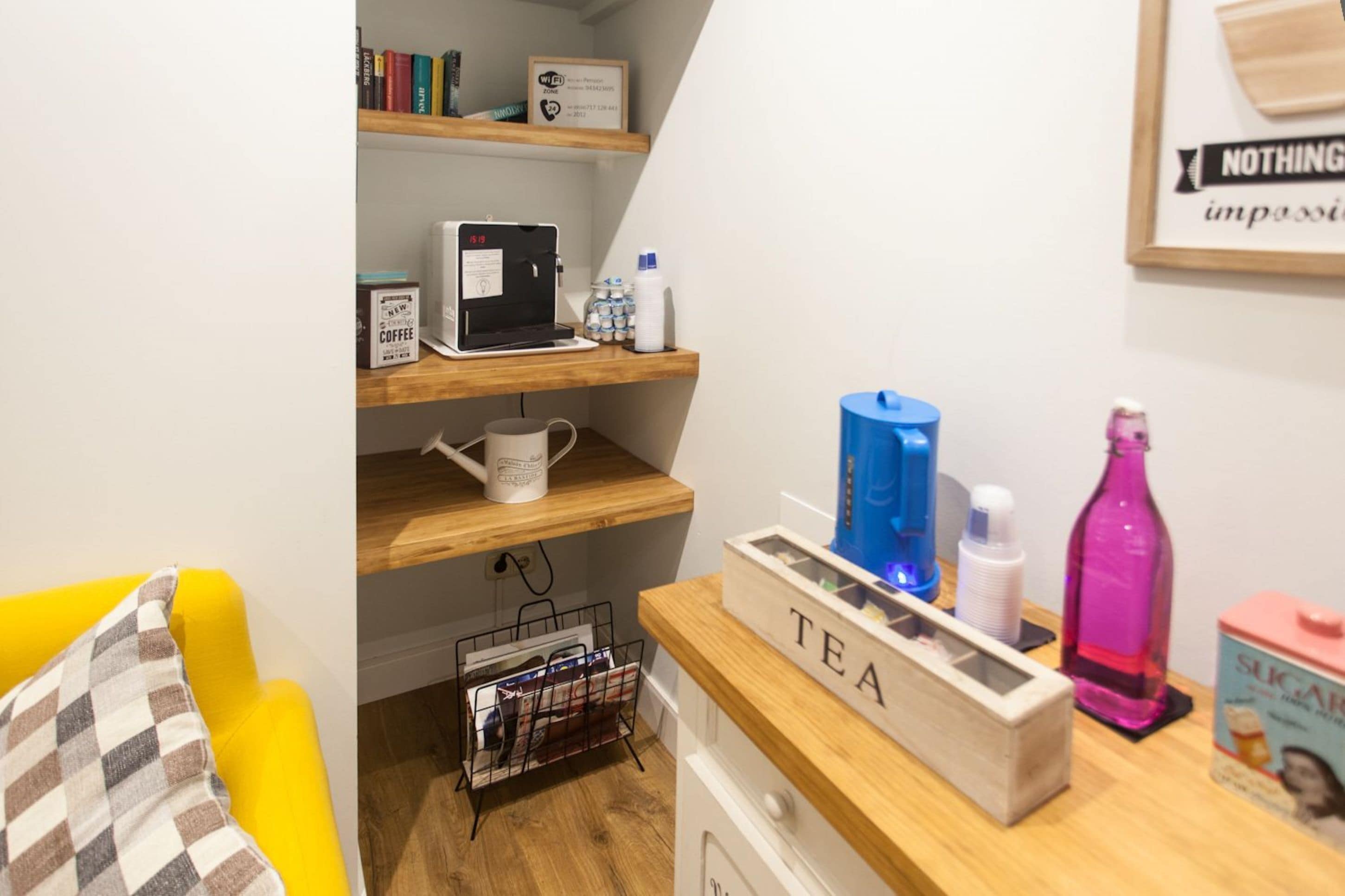 A cozy room with a wooden shelf holding books, a coffee machine, and a mug. There's a coffee machine on a lower shelf, a white mug, and a magazine rack. A wooden table with a 'TEA' sign and various items like a blue bottle, a pink bottle, and a box labeled 'SUGAR'.