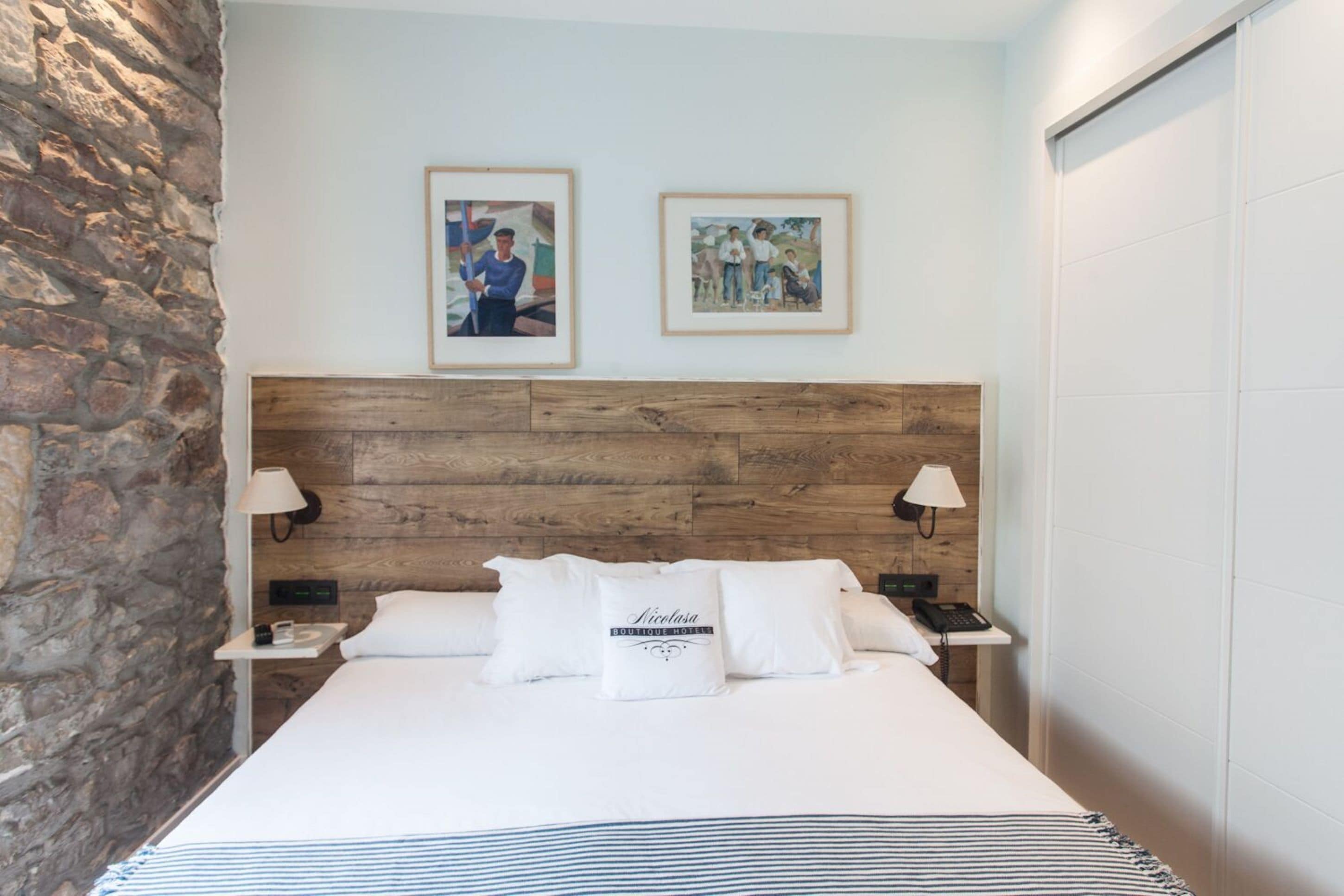 A hotel room with a stone wall, wooden headboard, two framed pictures, a bed with white bedding, and bedside lamps.
