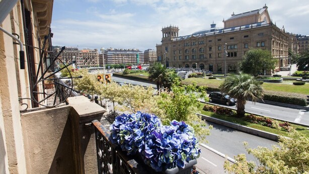 Un balcón con vistas a un paisaje urbano, que incluye un edificio grande de arquitectura clásica y un jardín bien cuidado.