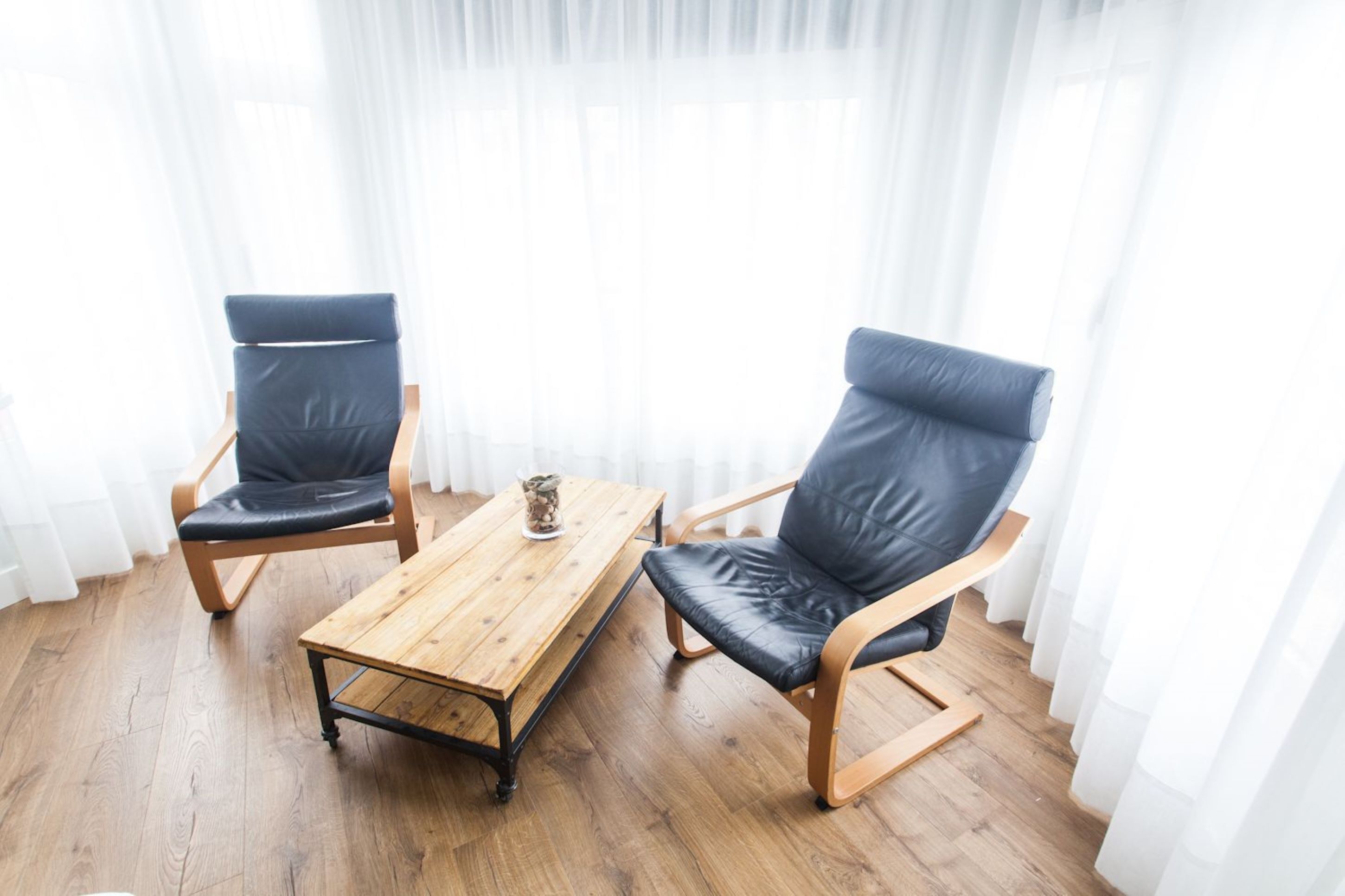 Two black leather chairs with wooden frames and a wooden coffee table in a room with wooden flooring and white curtains.