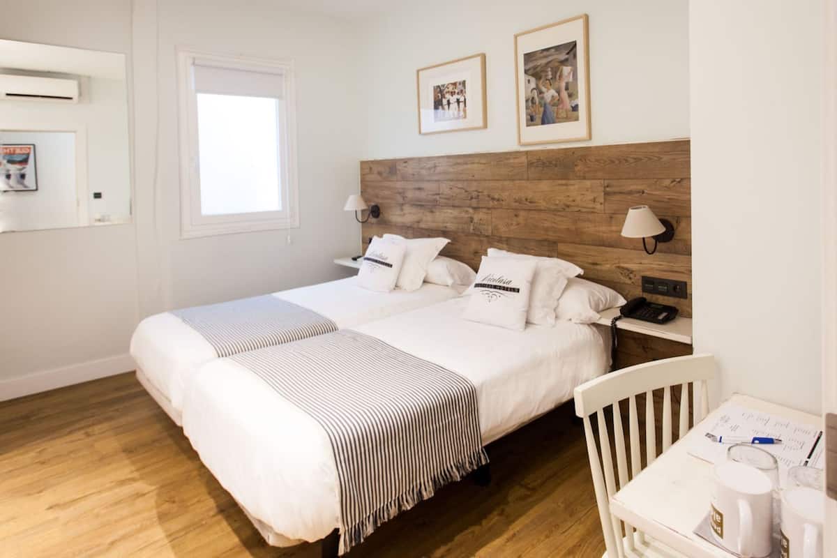 A double bed with a wooden headboard, two bedside tables with lamps, a small table with a chair, and a window.