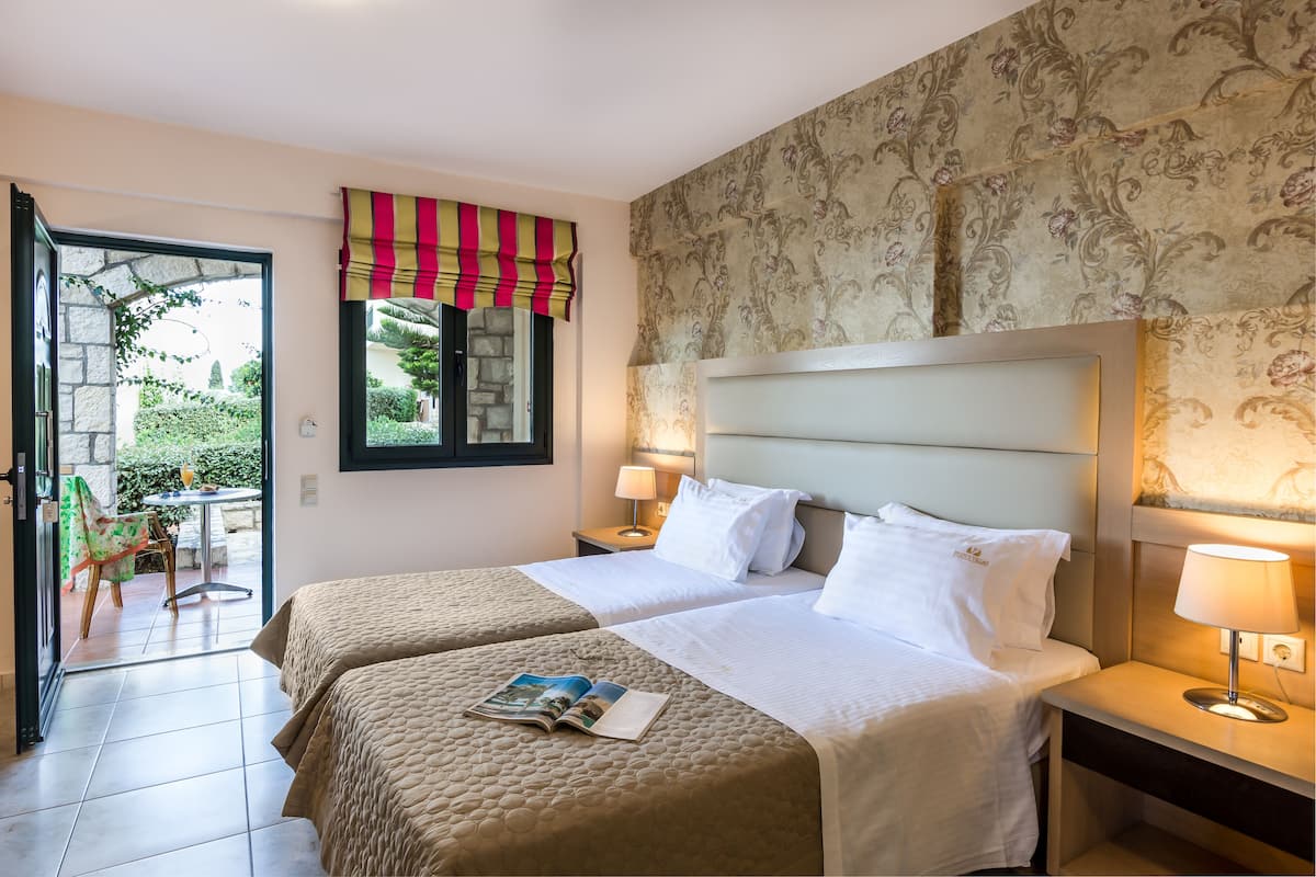 A hotel room with two beds, a window with a striped curtain, a lamp, and a view of the garden through the open door.