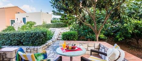 A patio with wicker furniture, a table with fruit and a drink, and a view of a building and greenery.