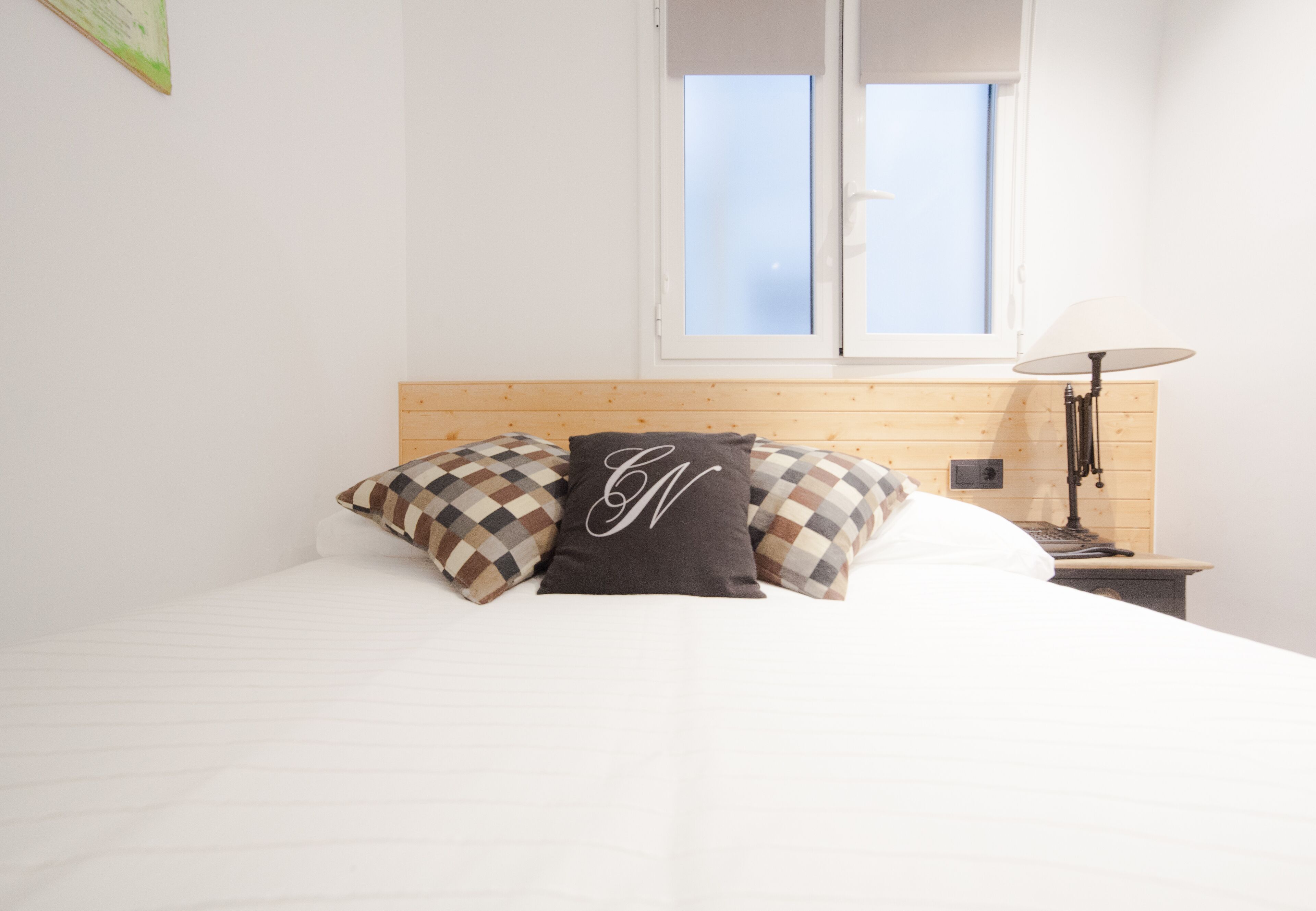 A neatly made bed with checkered pillows, a wooden headboard, a bedside lamp, and a window with blinds.