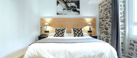 A bedroom with a wooden headboard, two bedside lamps, checkered pillows, a bed with a white duvet, and a stone wall.